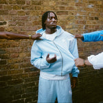 Man wearing pastel blue Five Questions Fleece zip-up hoodie against brick wall with hands pulling hoodie