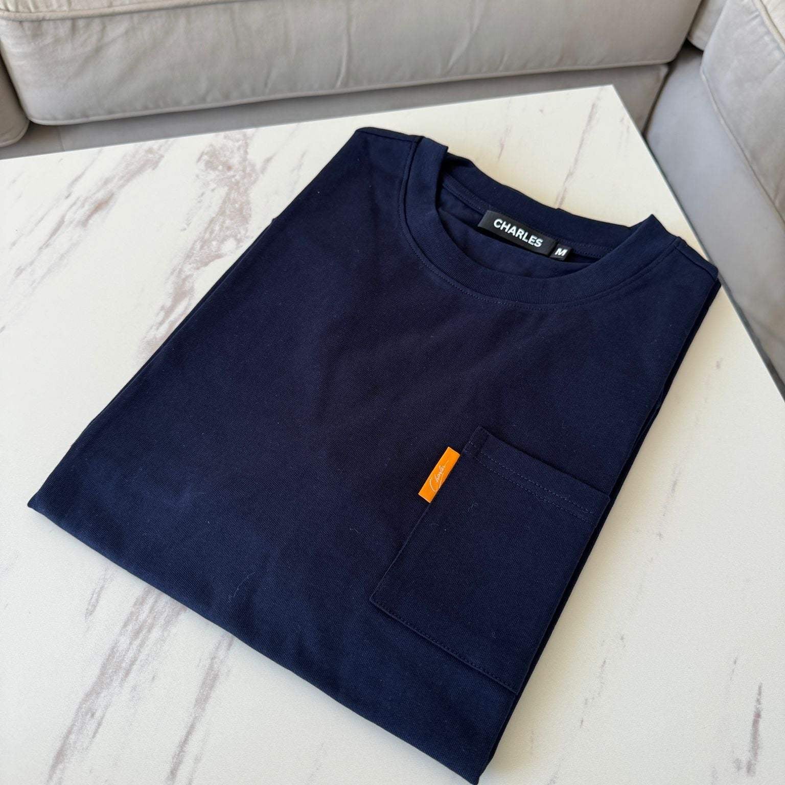 Studio By Charles navy pocket tee neatly folded on white marble table next to beige sofa