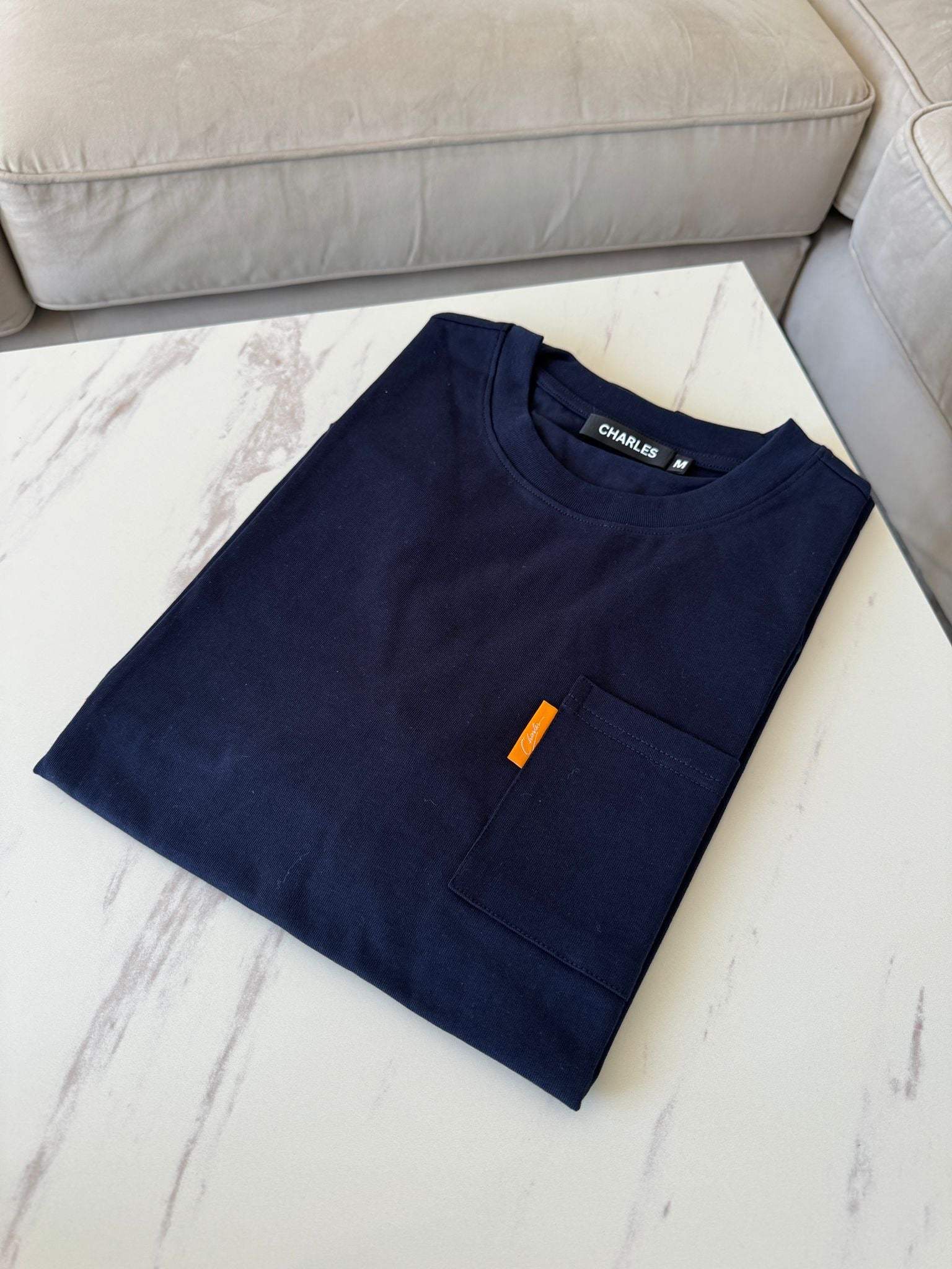 Studio By Charles navy pocket tee neatly folded on white marble table next to beige sofa