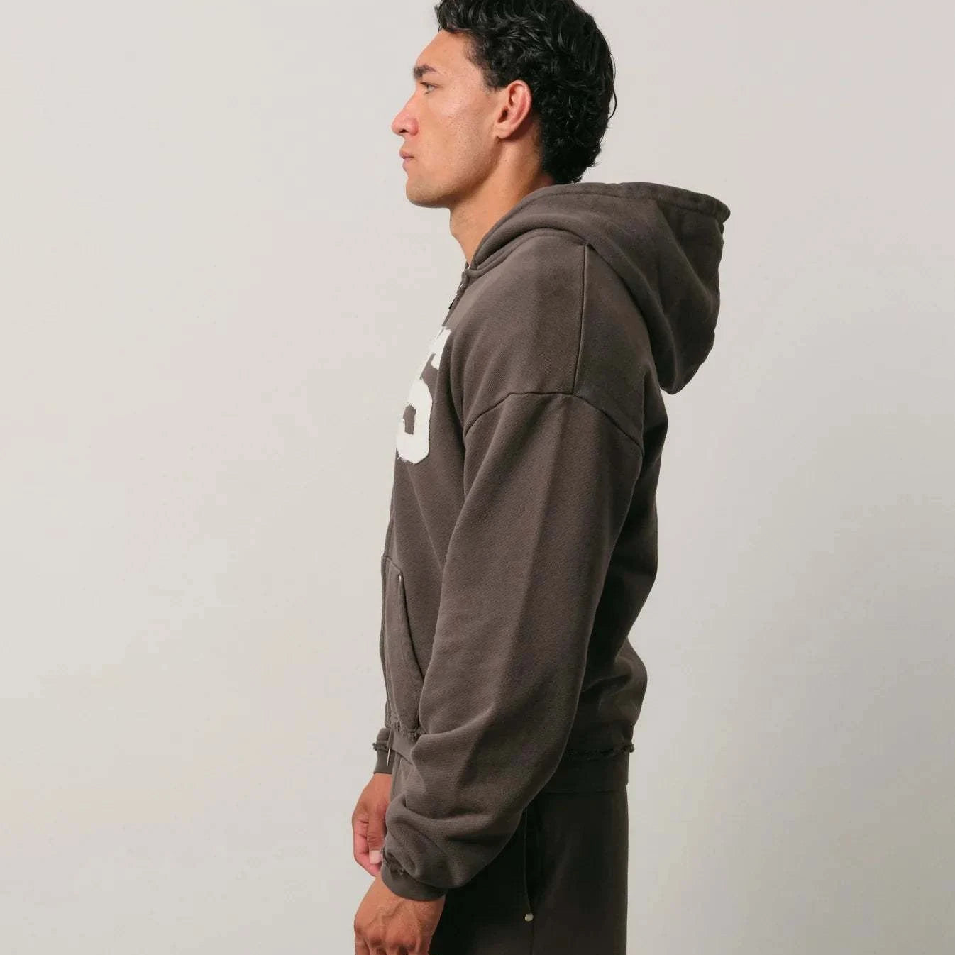 Side view of a man wearing a dark brown Natural Art Studio NAS appliqué hoodie with a hood and dropped shoulder design.