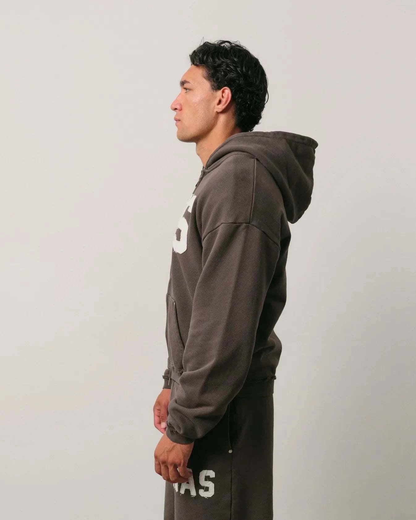 Side view of a man wearing a dark brown Natural Art Studio NAS appliqué hoodie with a hood and dropped shoulder design.