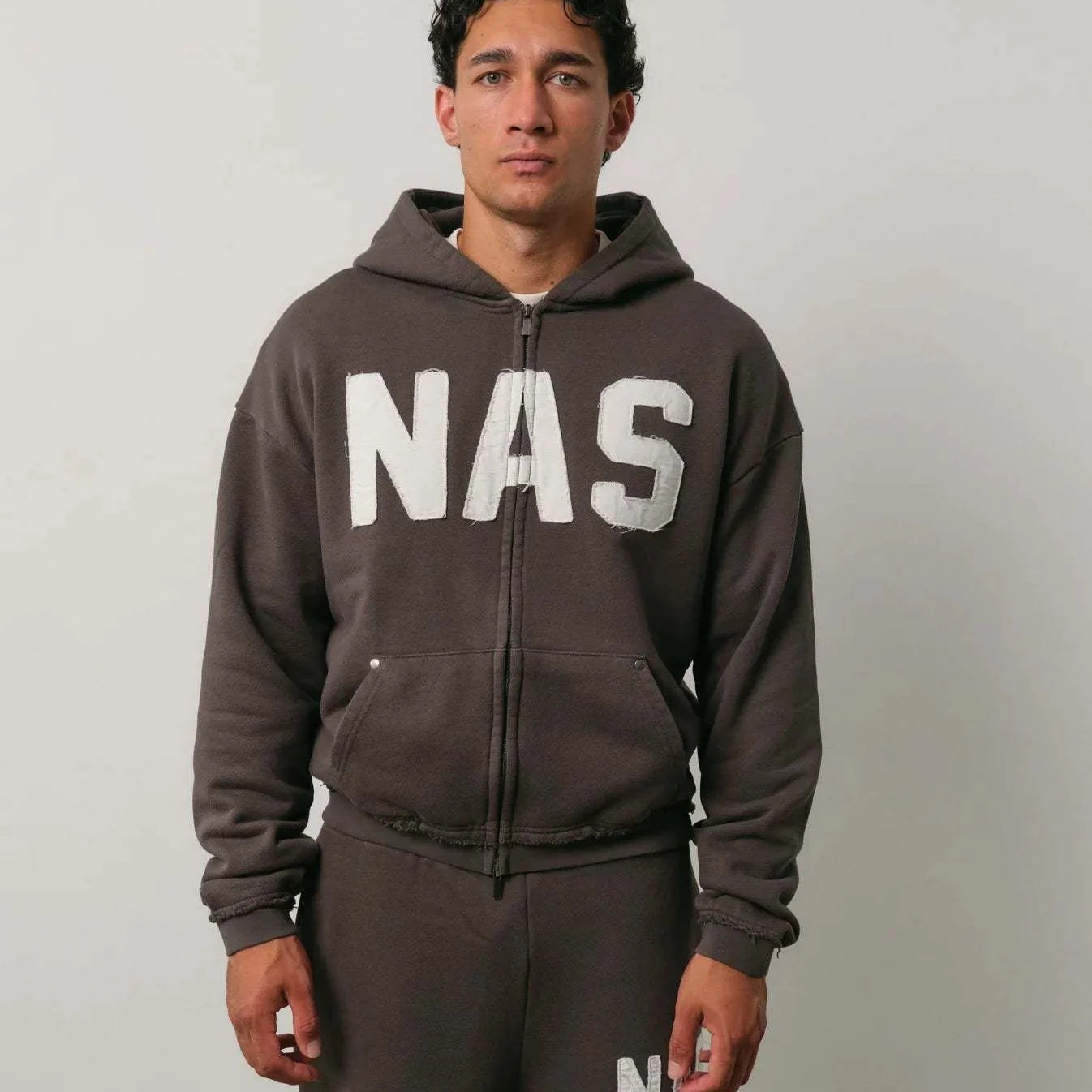 Man wearing Natural Art Studio NAS appliqué hoodie in dark color with white NAS logo and matching pants