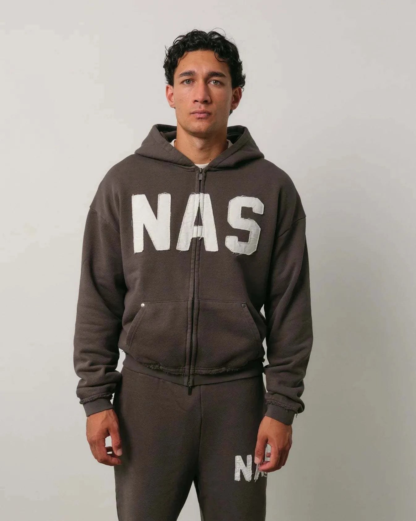 Man wearing Natural Art Studio NAS appliqué hoodie in dark color with white NAS logo and matching pants