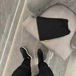 Studio By Charles black trackpants folded on gray chair with person wearing black trackpants and sneakers on gray tiled floor