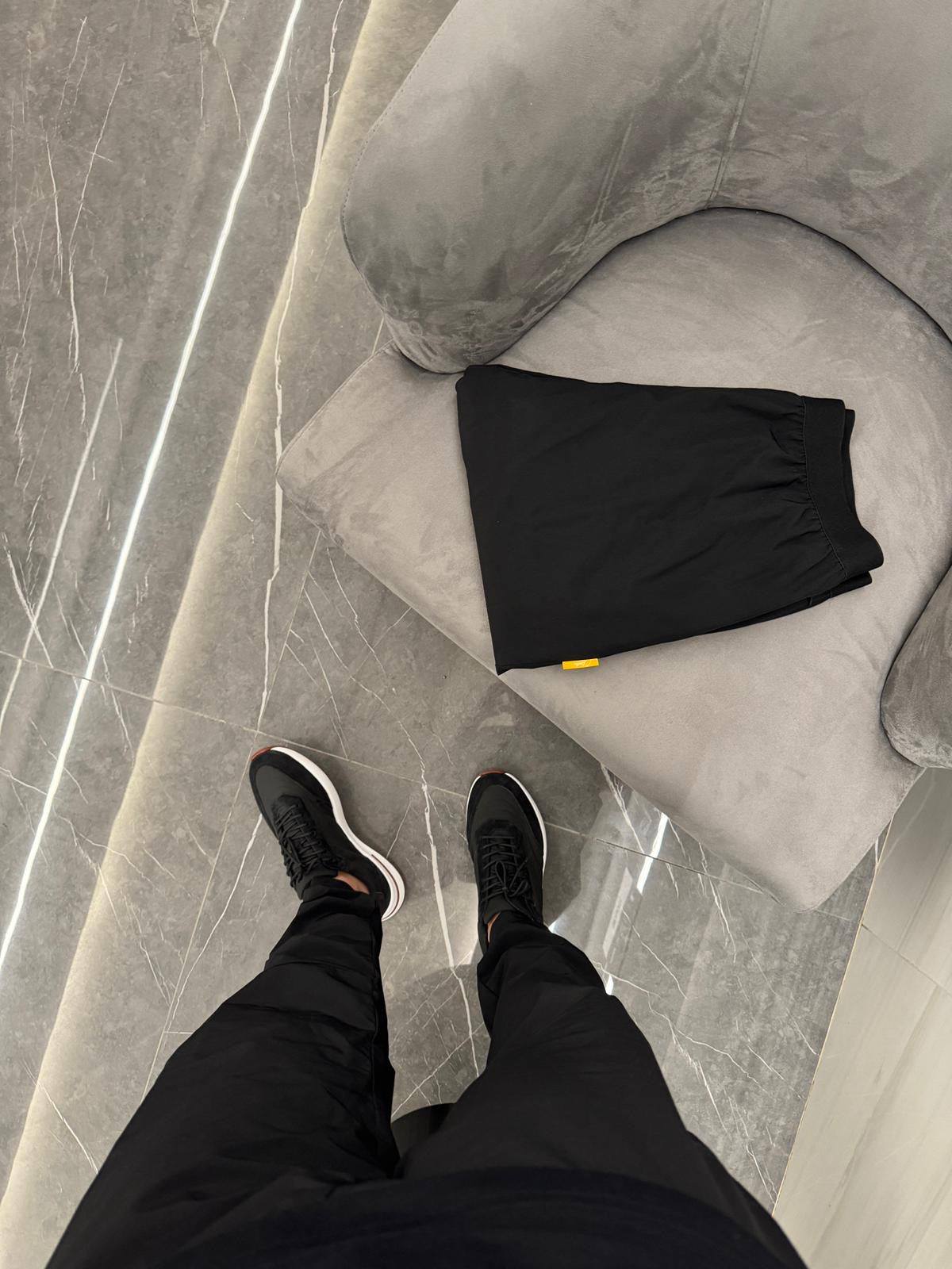 Studio By Charles black trackpants folded on gray chair with person wearing black trackpants and sneakers on gray tiled floor