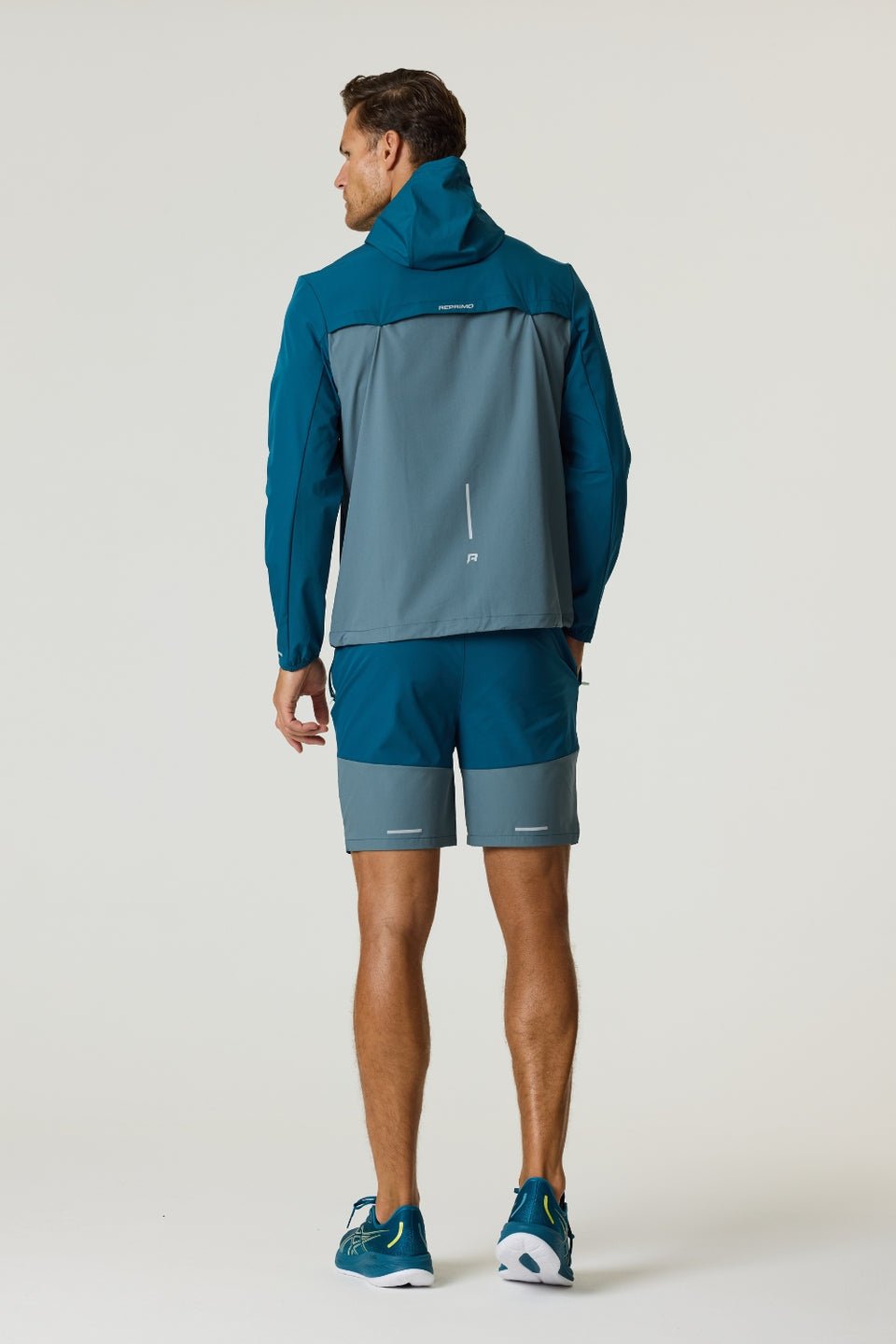 Man wearing Reprimo Flight Windbreaker in tidal teal and blue steel from the back, showcasing lightweight weather-resistant jacket with hood and zippered pockets.