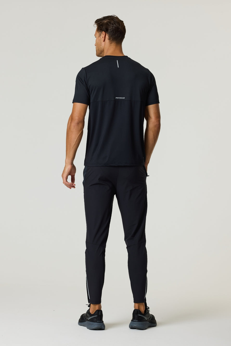 Rear view of a man wearing a black Reprimo Flight T-Shirt with reflective strips and athletic fit, paired with black workout pants.