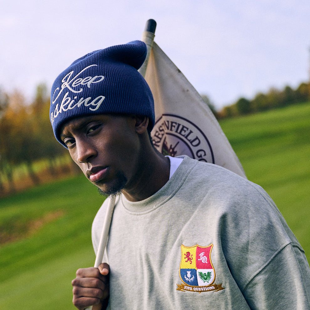 Man wearing navy Five Questions Keep Asking slouch beanie outdoors with a flag and gray sweatshirt