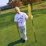 Young man wearing white Five Questions Western Tee standing on golf course holding flagstick