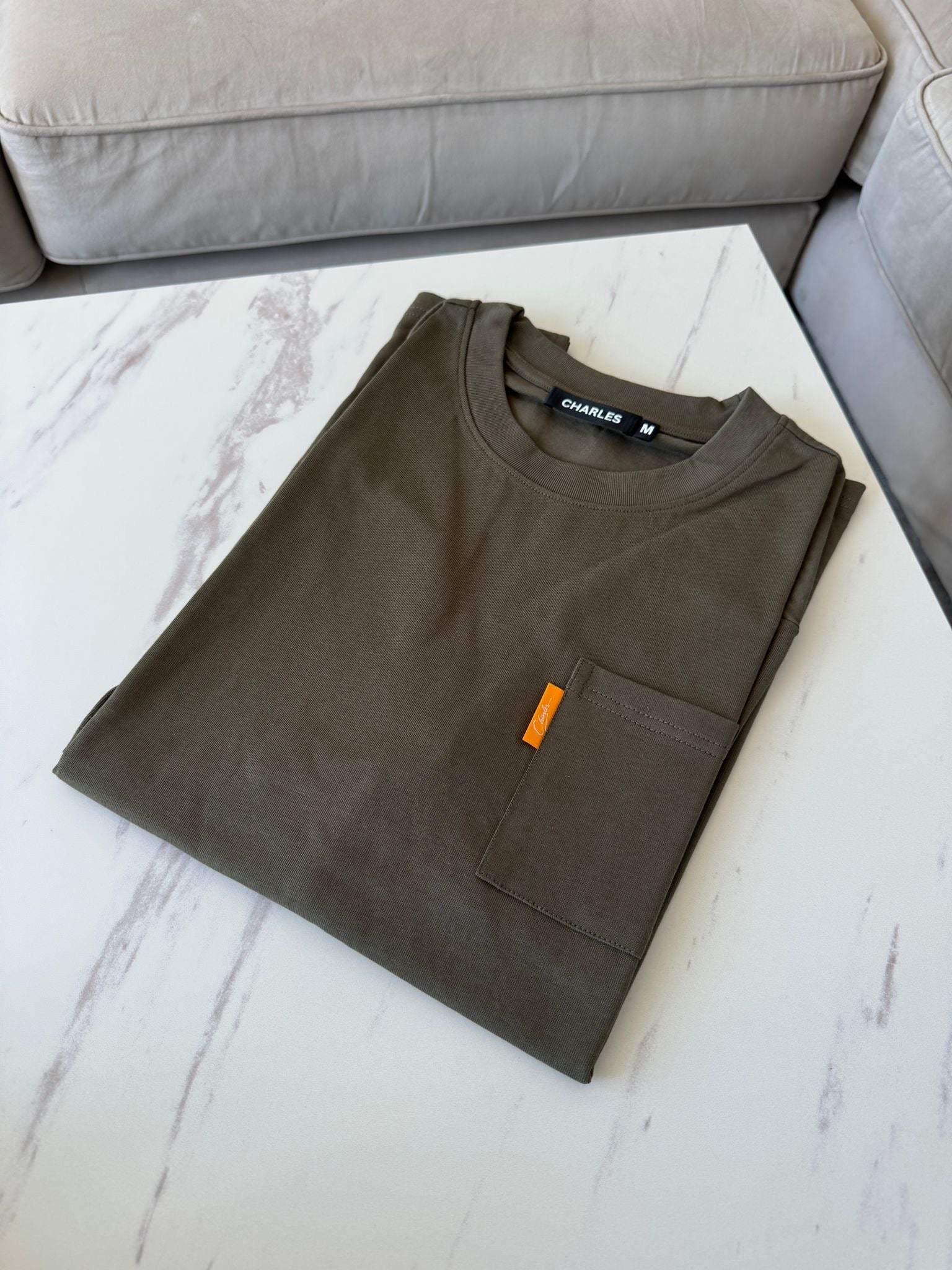 Folded khaki Studio By Charles pocket tee on white marble surface