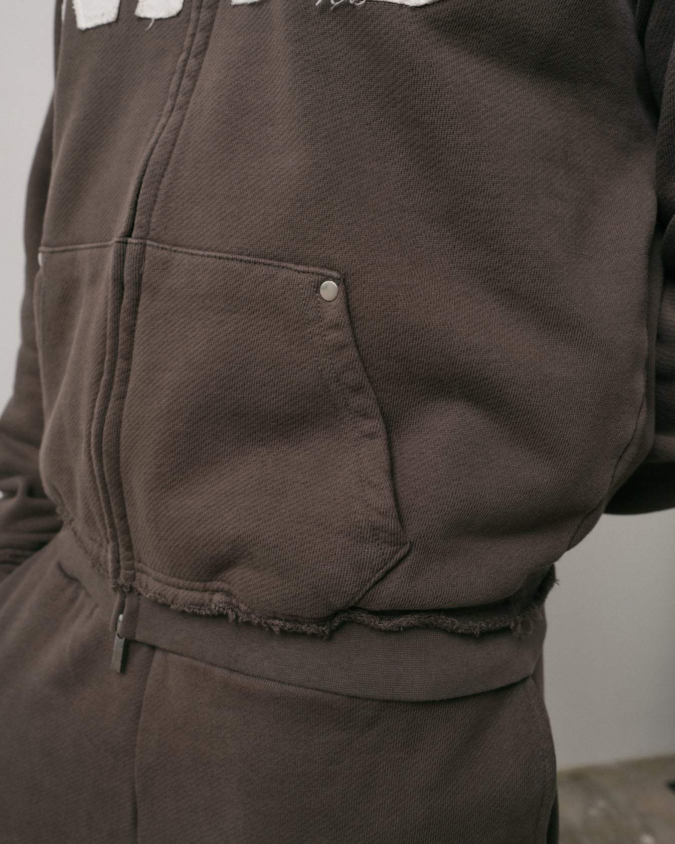 Close-up of brown Natural Art Studio NAS Appliqué Hoodie showing raw seams on cuffs and metal rivet on pocket detail