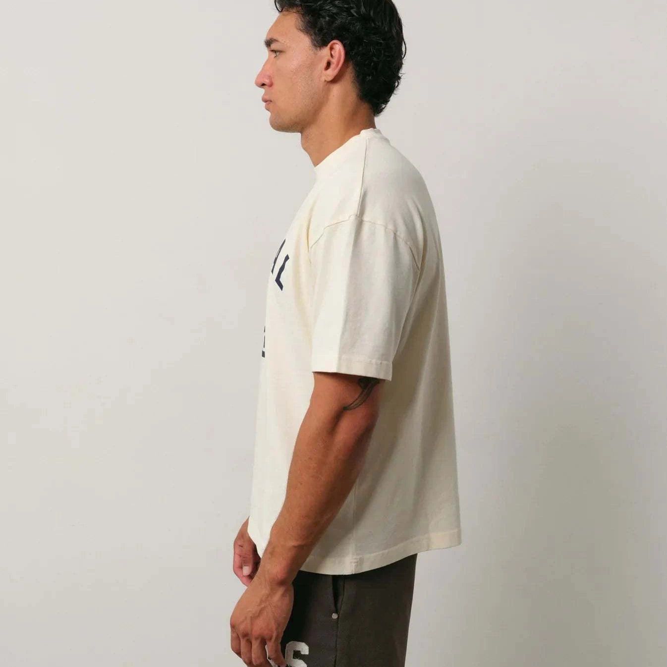 Natural Art Studio Varsity Tee 100% cotton boxy fit oversized drop shoulder cream shirt with front and back screen print logo worn by male model side view
