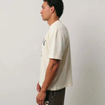 Natural Art Studio Varsity Tee 100% cotton boxy fit oversized drop shoulder cream shirt with front and back screen print logo worn by male model side view