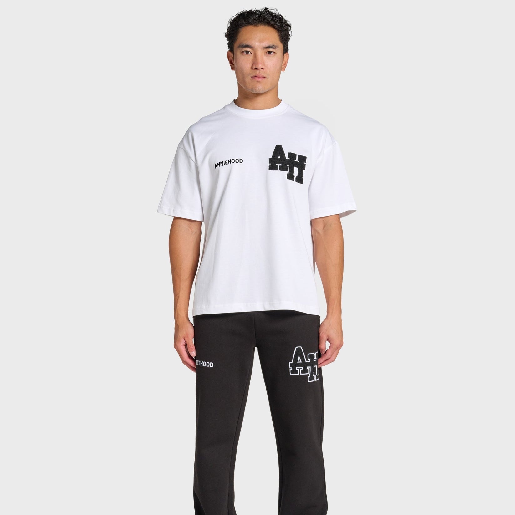 Male model wearing white ANNIE HOOD college t-shirt with black logo and black pants with matching ANNIE HOOD branding