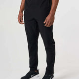 Man wearing Cova Daily Trousers black slim-fit pants with black and white sneakers against a plain background