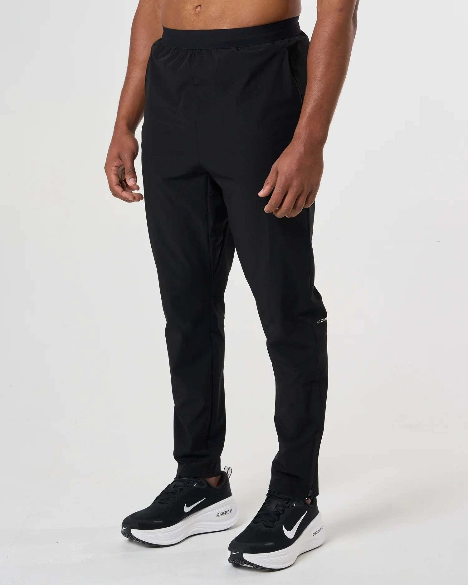 Man wearing Cova Daily Trousers black slim-fit pants with black and white sneakers against a plain background