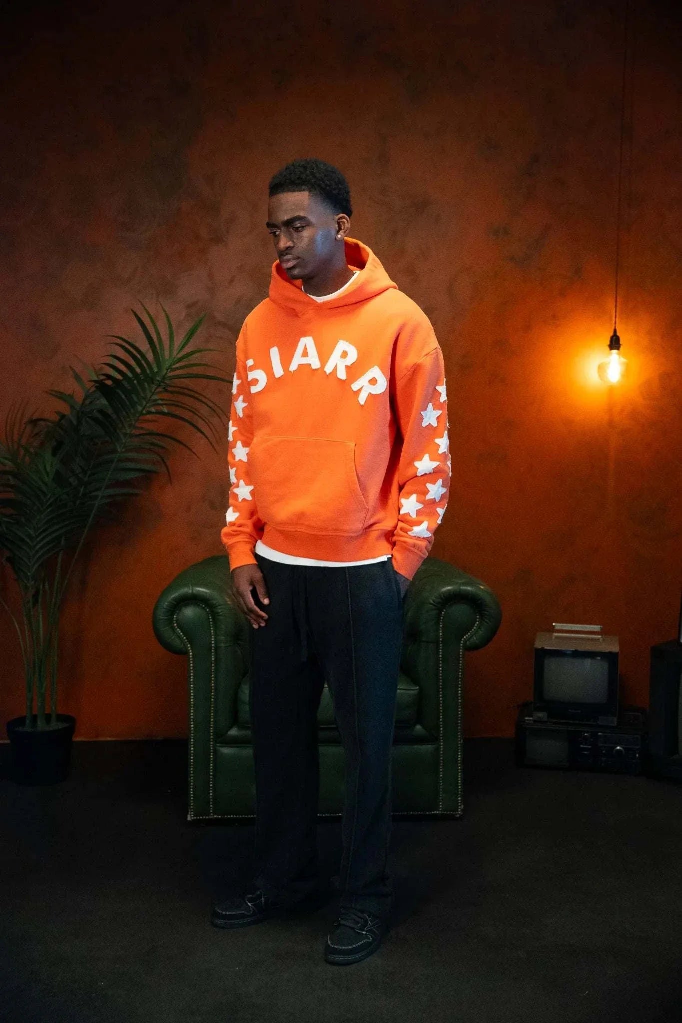 Man wearing orange Siarr Applique Hoodie with white stars on sleeves and kangaroo pocket