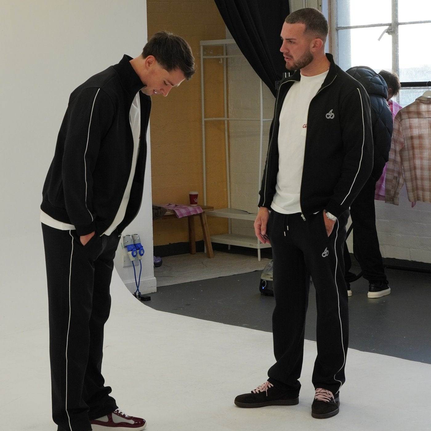 Men wearing Cherry Location black tracksuit bottoms with white side stripes in a casual indoor setting