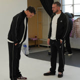 Men wearing Cherry Location black tracksuit bottoms with white side stripes in a casual indoor setting