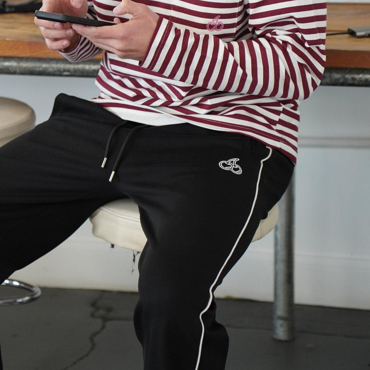 Man wearing Cherry Location black tracksuit bottoms with white side piping and logo detail sitting on a stool indoors