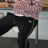 Man wearing Cherry Location black tracksuit bottoms with white side piping and logo detail sitting on a stool indoors