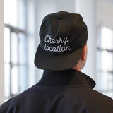 Cherry Location Black Cap worn on a person's head showing embroidered white text