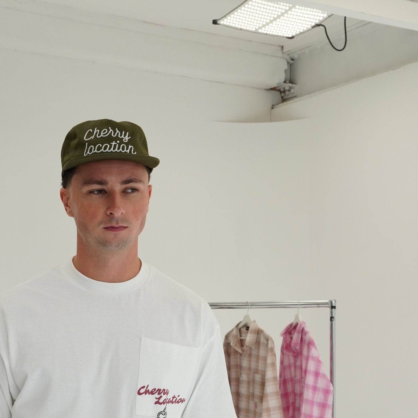 Man wearing Cherry Location Green Cap and white shirt in minimal room with clothing rack