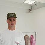 Man wearing Cherry Location Green Cap and white shirt in minimal room with clothing rack