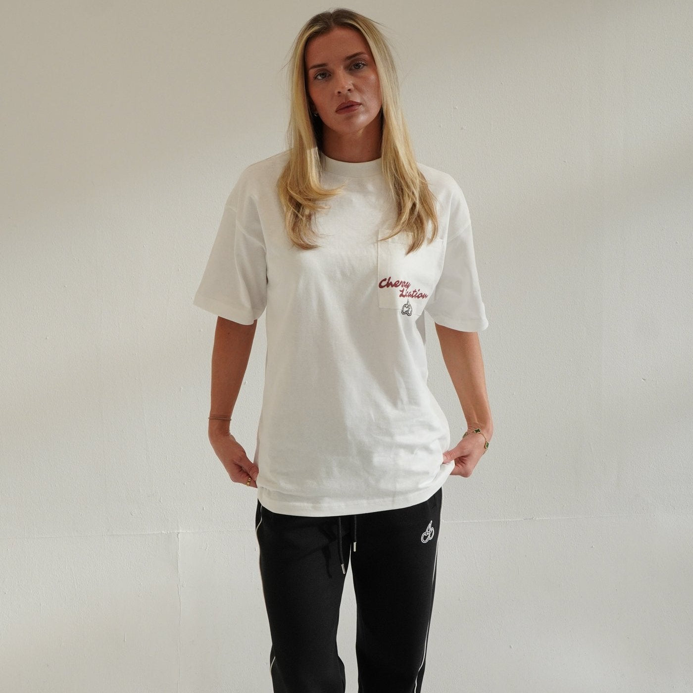Woman wearing Cherry Location white t-shirt with red text and black pants standing against plain wall
