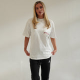 Woman wearing Cherry Location white t-shirt with red text and black pants standing against plain wall