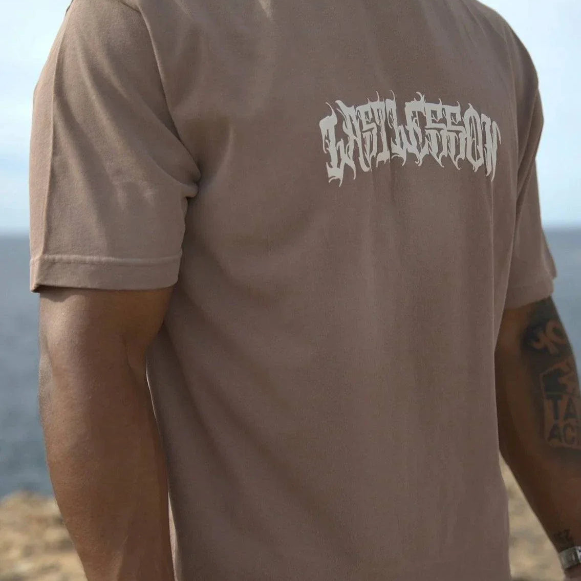 Last Lesson brown script t-shirt worn by man standing outdoors