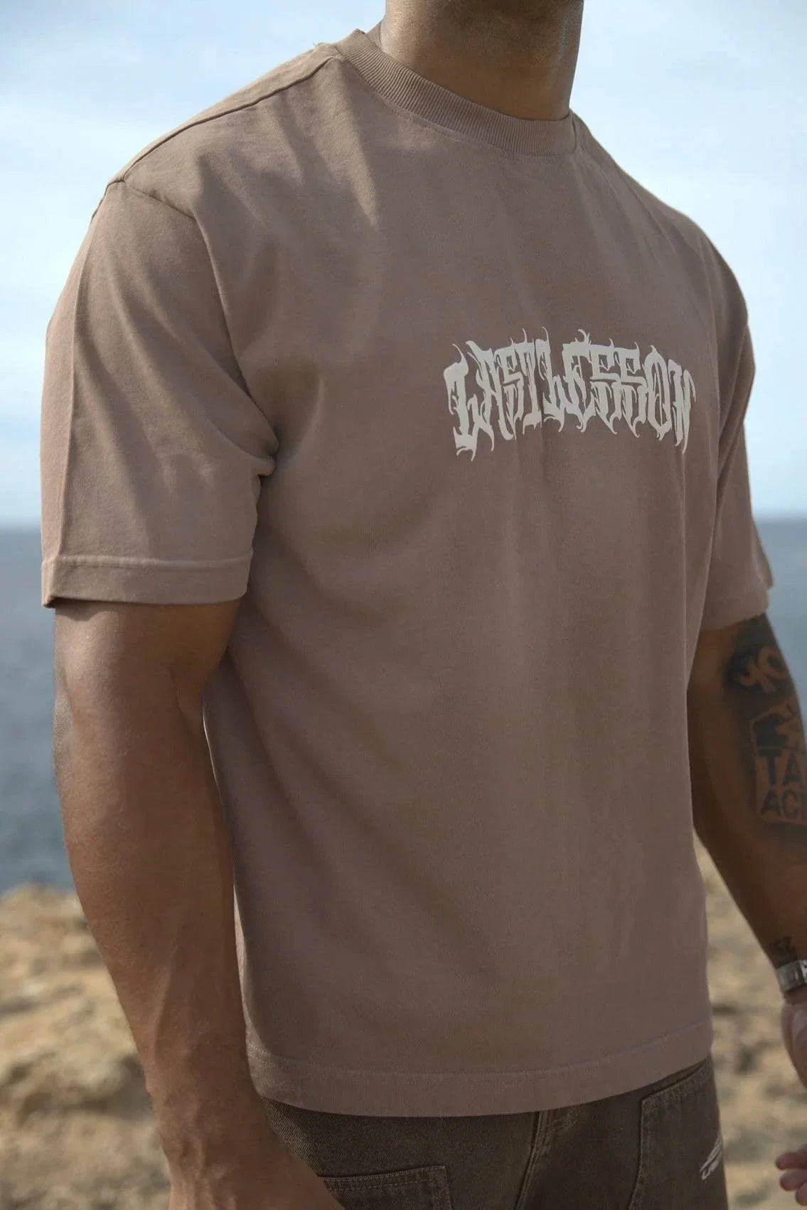 Last Lesson brown script t-shirt worn by man standing outdoors
