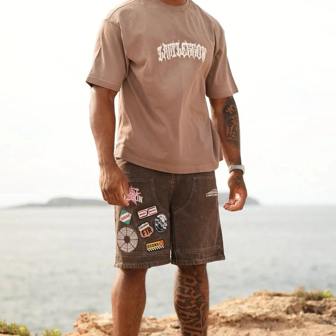 Man wearing Last Lesson brown script t-shirt with patched brown shorts and white sneakers outdoors