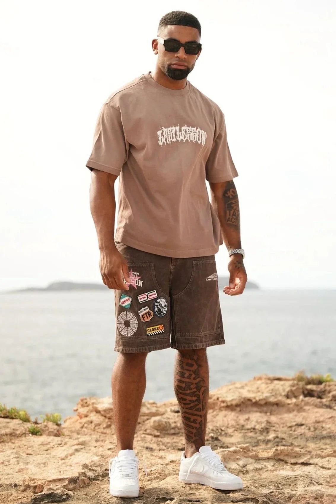 Man wearing Last Lesson brown script t-shirt with patched brown shorts and white sneakers outdoors