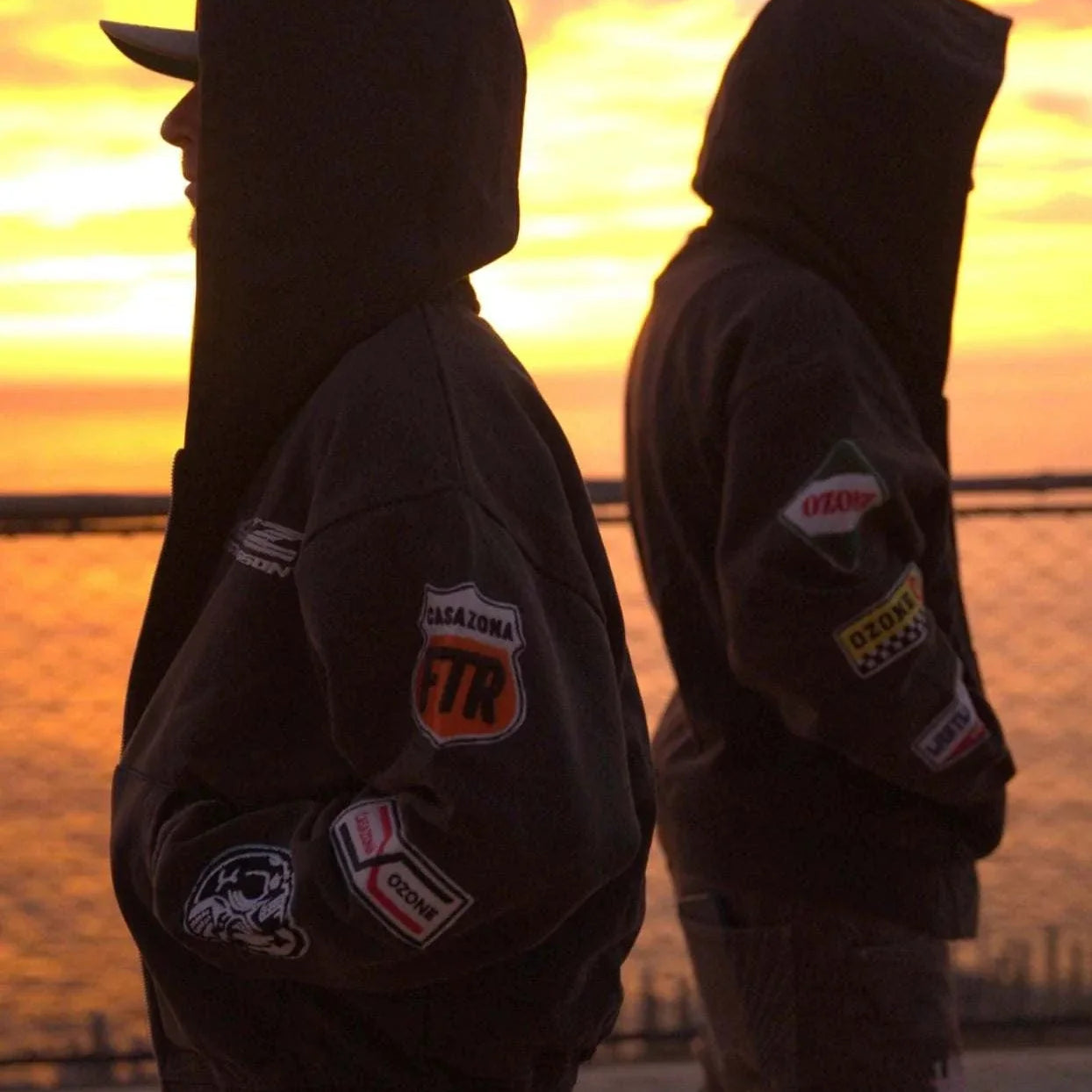 Two people wearing Last Lesson Badged Casazøna Hoodies standing back-to-back at sunset