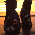 Two people wearing Last Lesson Badged Casazøna Hoodies standing back-to-back at sunset