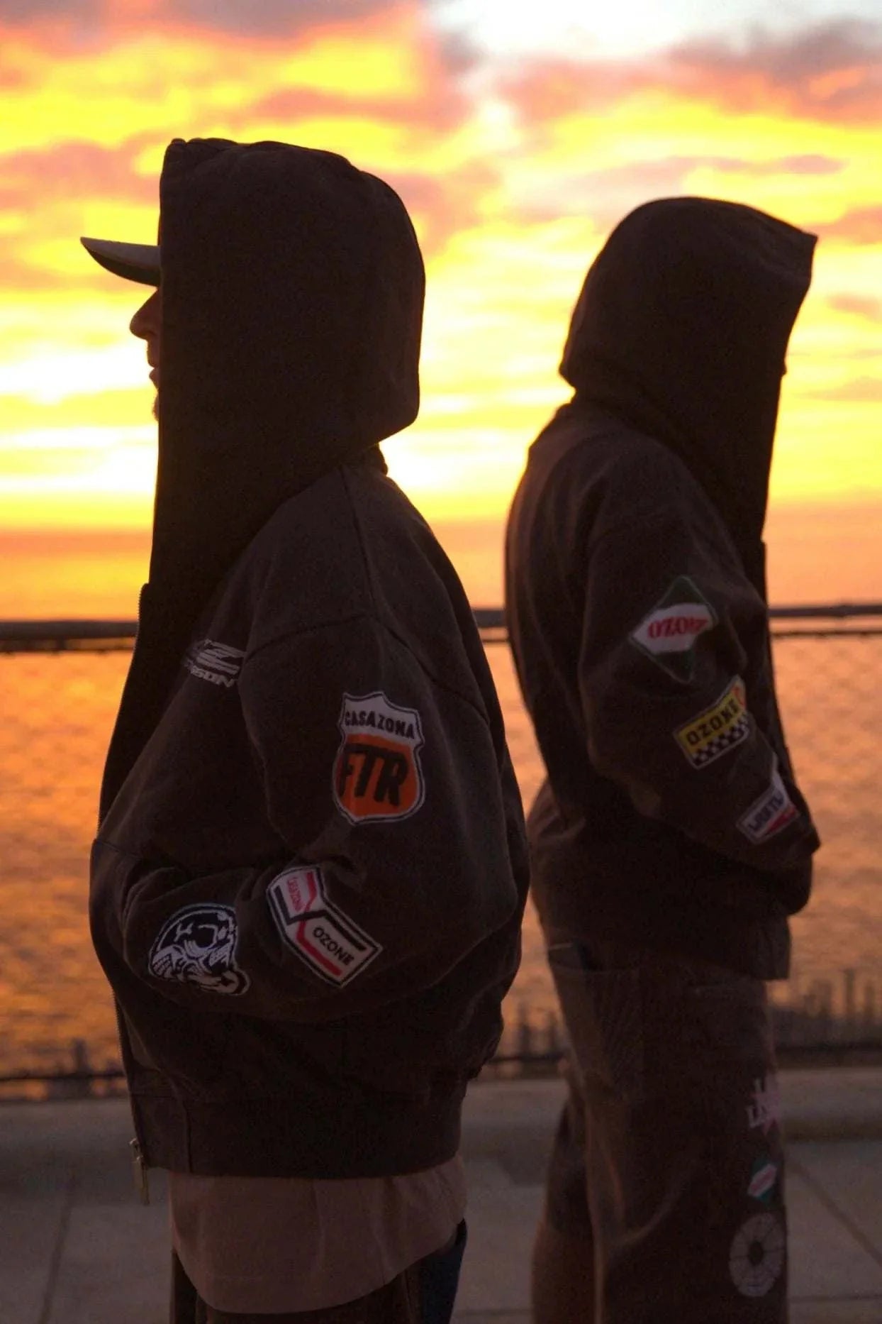 Two people wearing Last Lesson Badged Casazøna Hoodies standing back-to-back at sunset