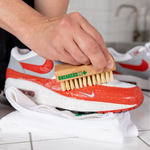 Hand using SneakersER Premium Cleaning Duo Kit brush to clean white and red Nike sneakers on white towel
