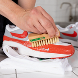 Hand using SneakersER Premium Cleaning Duo Kit brush to clean white and red Nike sneakers on white towel