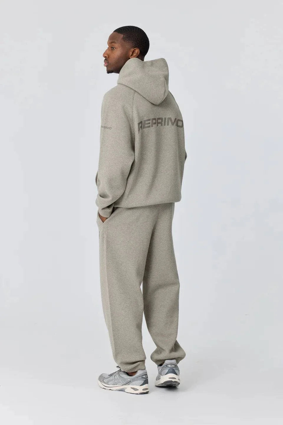 Man wearing grey Reprimo Collective Hoodie with matching pants and sneakers, showing back design and reflective logo.