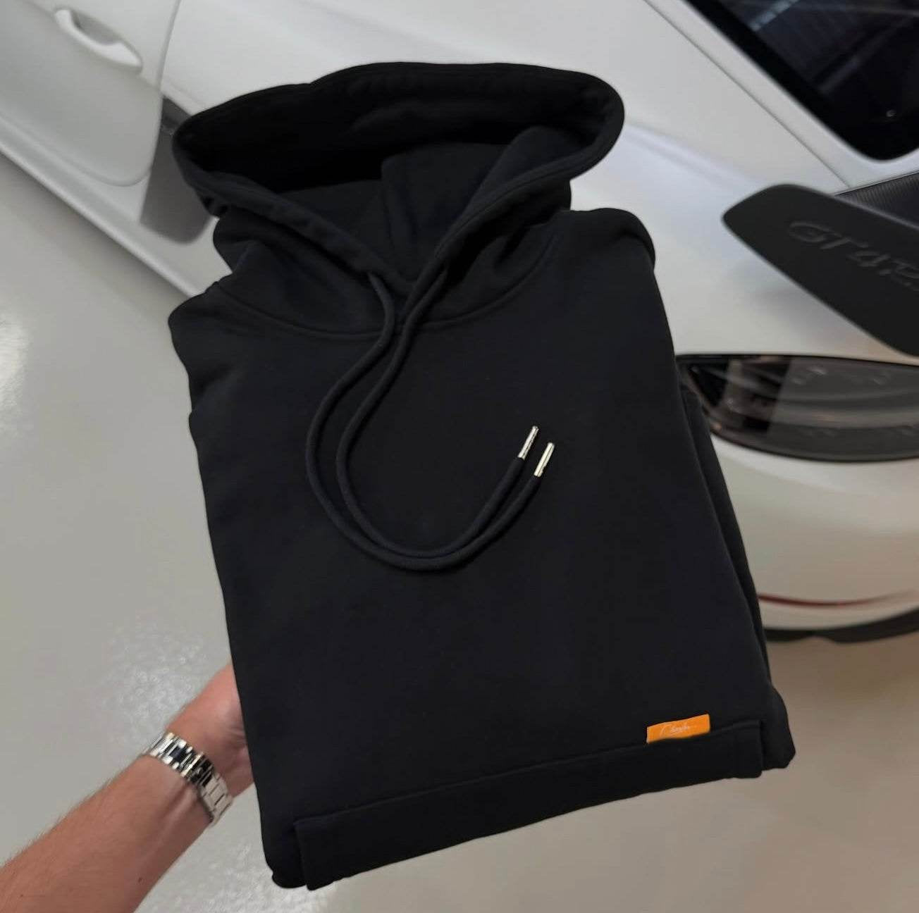 Folded black Studio By Charles Hoodie with drawstrings and small orange tag held above white car background