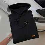 Folded black Studio By Charles Hoodie with drawstrings and small orange tag held above white car background