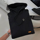 Folded black Studio By Charles Hoodie with drawstrings and small orange tag held above white car background