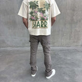 Back view of a person wearing a cream oversized Siarr Palm Tee with palm tree graphic and "SIARR" text, paired with grey pants and white shoes.