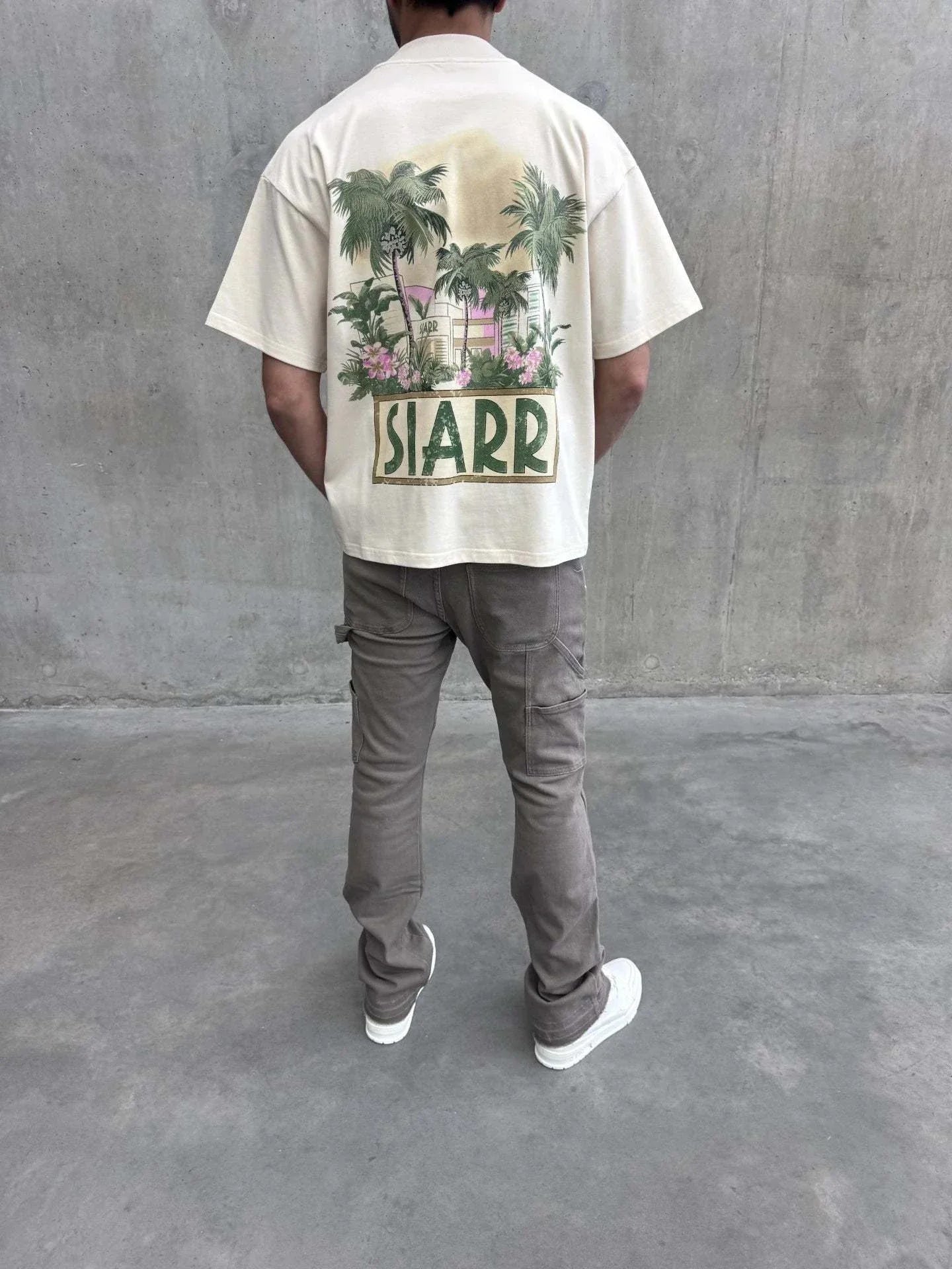 Back view of a person wearing a cream oversized Siarr Palm Tee with palm tree graphic and "SIARR" text, paired with grey pants and white shoes.
